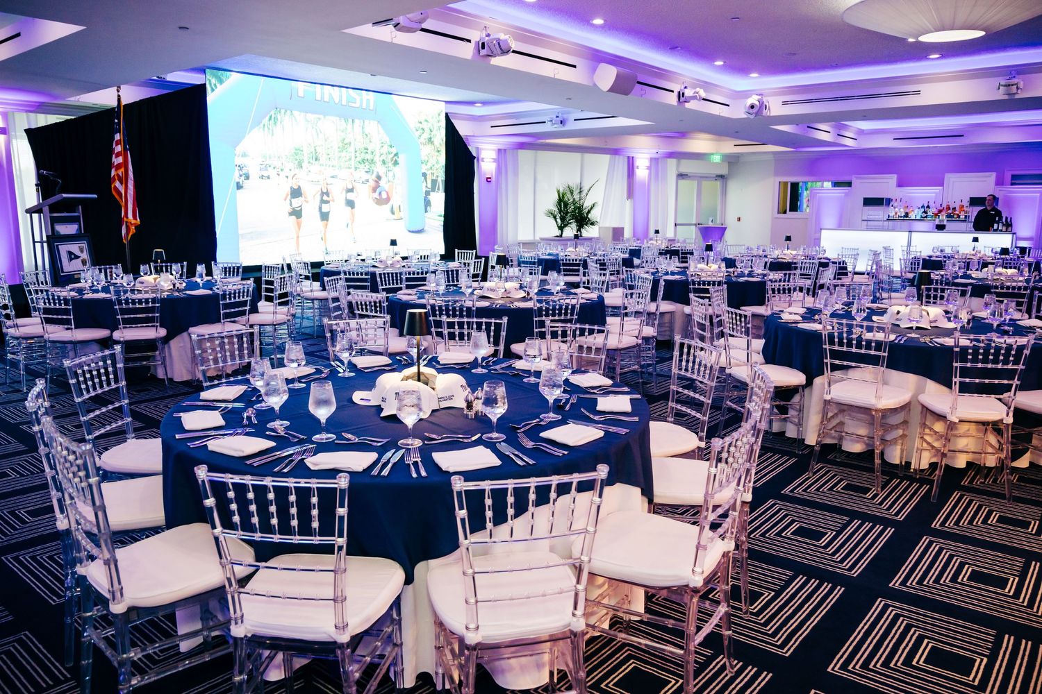 Elegant banquet hall with navy blue round tables, crystal chiavari chairs, and a large projection screen for events.