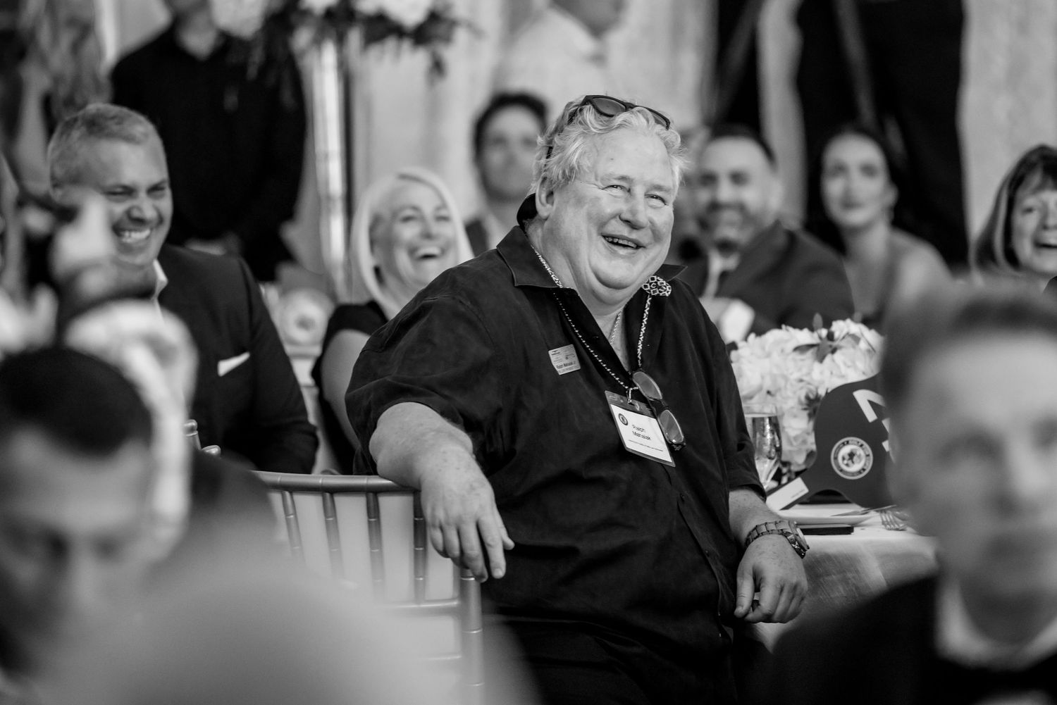 A cheerful older man laughs at an event, wearing a black shirt with a name badge, surrounded by guests in a elegant venue.