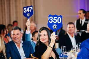 Guests at a charity auction gala raise blue numbered bidding paddles 347 and 350 while seated at an elegant dinner event.