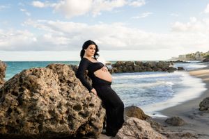 Maternity photo of woman in black dress posing on beach rocks by ocean.