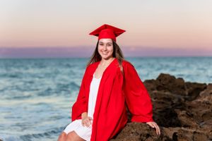 Graduate in red cap and gown poses on rocky beach at sunset.