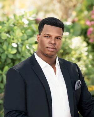 Professional headshot of person in black suit with white shirt and pocket square.