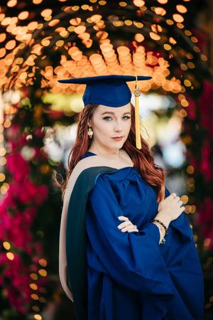 Graduate in blue cap and gown poses with bokeh lights and flowers in background.