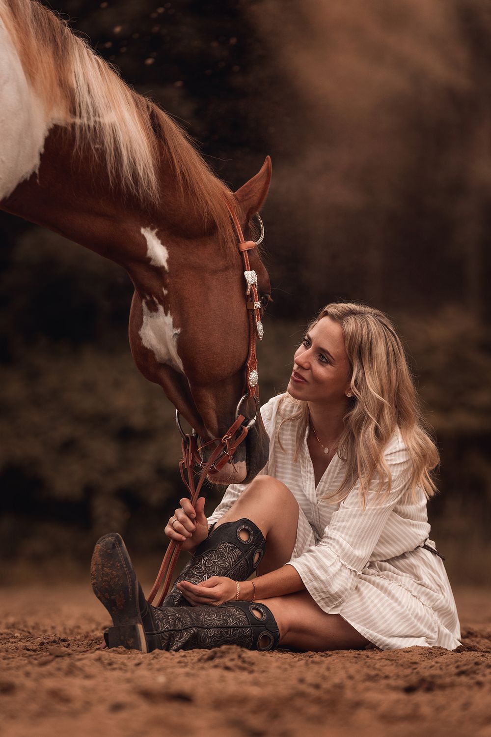 Blonde Frau in weißem Kleid sitzt auf Sandboden und kuschlet mi ihrem gescheckten Pferd