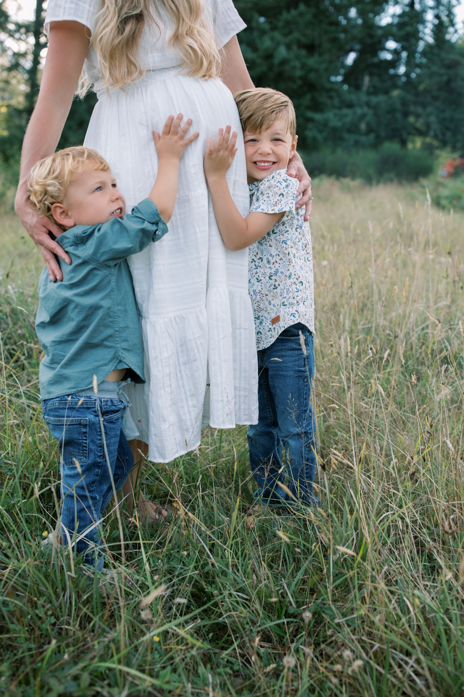 Two young boys hugging their pregnant mother's belly in a grassy field.