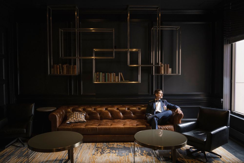 A military groom sits in a dark elegant room with brown leather sofa, modern gold shelving, and natural light pouring in through a window.