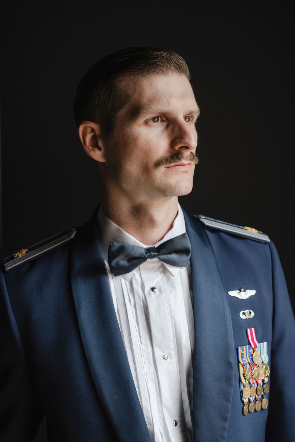 Military officer groom in dress blue uniform with bow tie and medals poses against dark background for portrait.