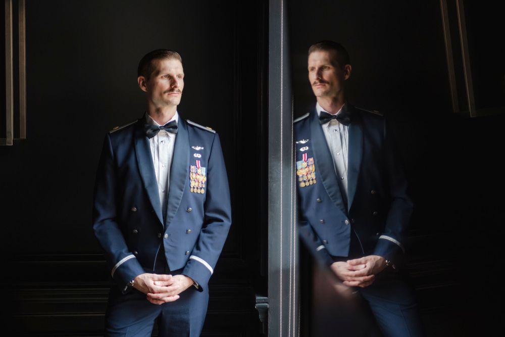 Military groom in formal blue suit and bowtie is reflected in mirror against dark background.