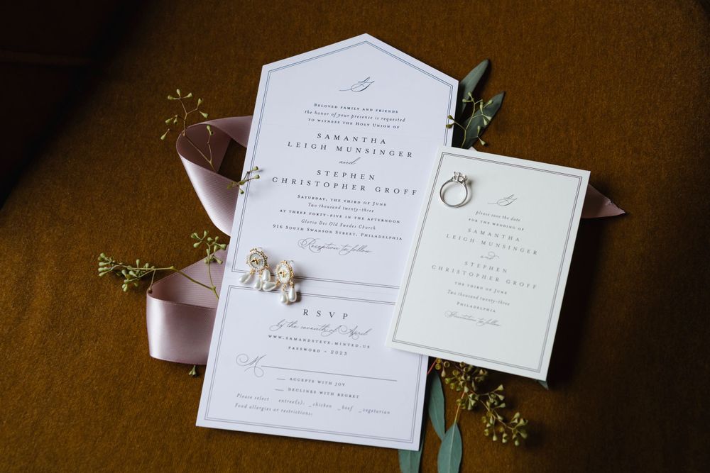 Elegant white wedding invitations with silver text displayed alongside rings and delicate pink ribbon on brown surface.