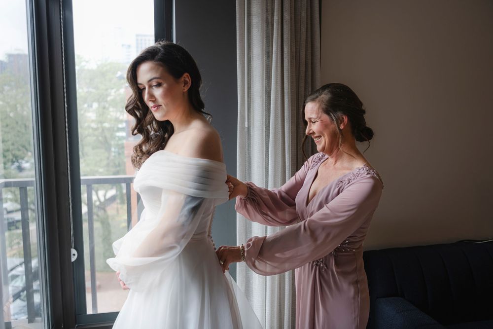 A bride in a white off-shoulder wedding dress gets help with final preparations from her mom near a window in natural light.