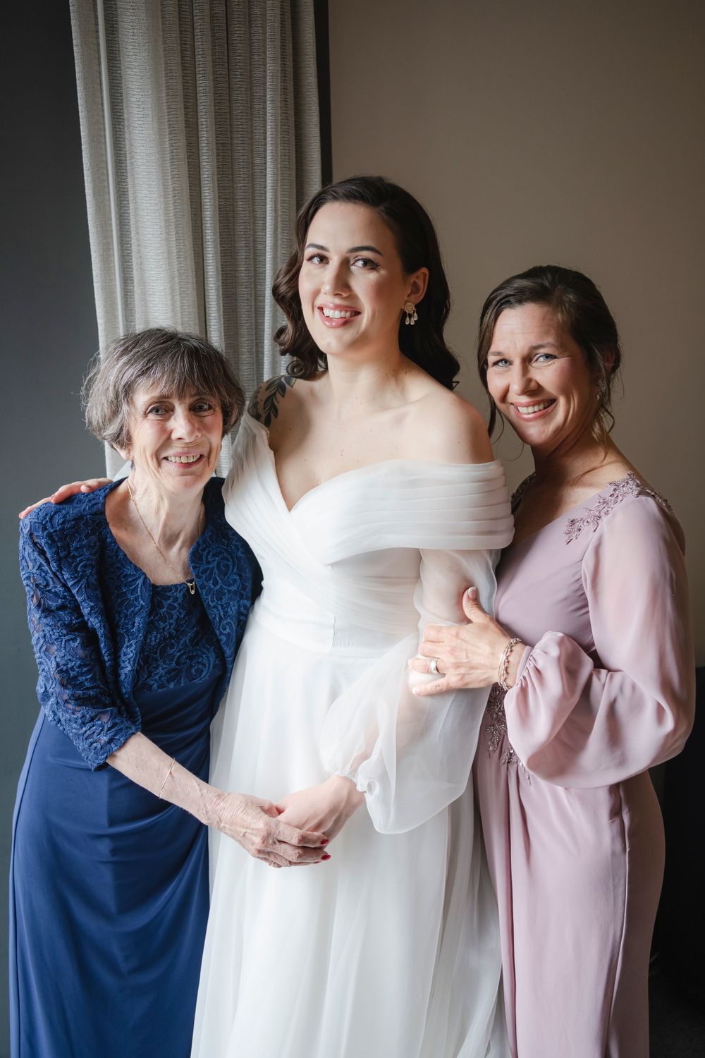 Three generations of women pose together by a window, with the central figure wearing a white off-shoulder wedding dress.