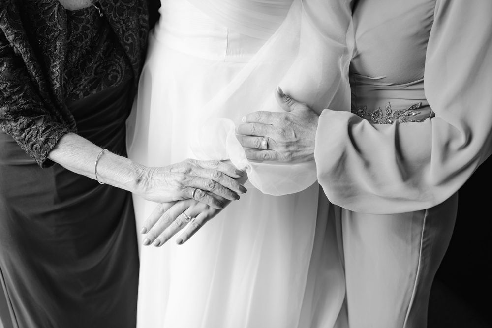 Black and white photo showing hands held together with delicate rings and wedding attire details.