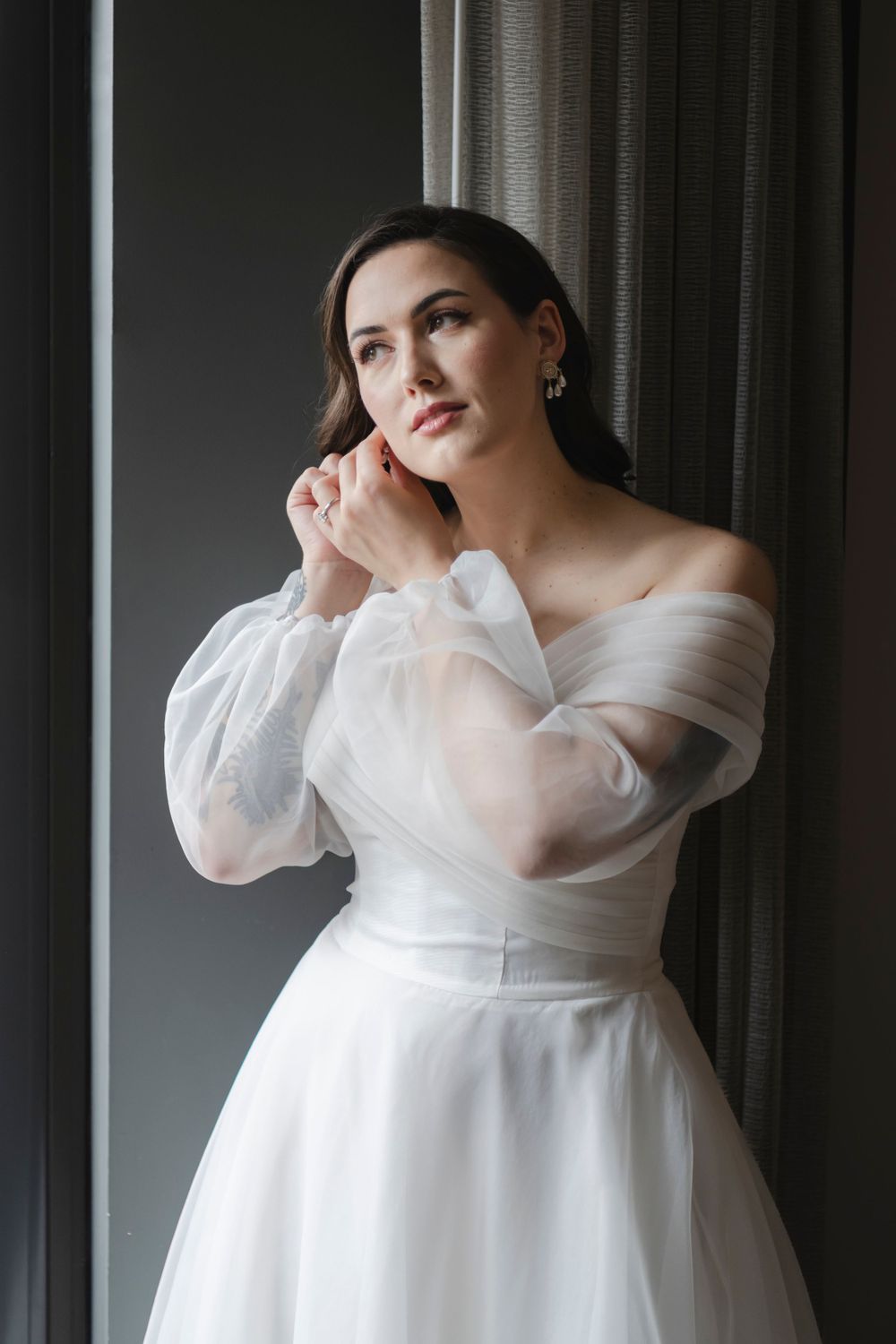Elegant bride in off-shoulder white tulle wedding gown with sheer sleeves poses by window in soft natural light.