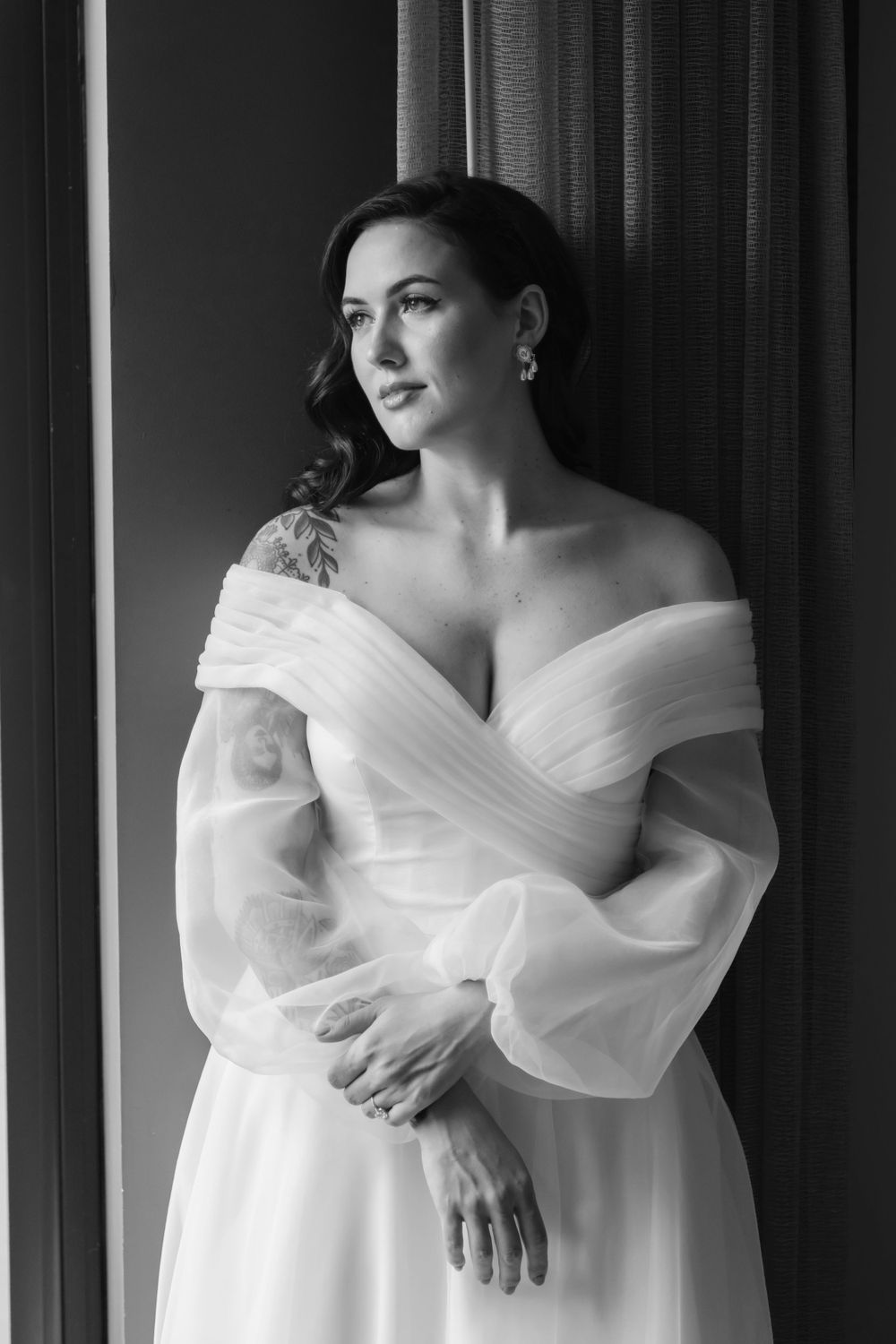 A bride in an elegant off-shoulder white wedding gown with sheer bishop sleeves poses by a window in black and white.