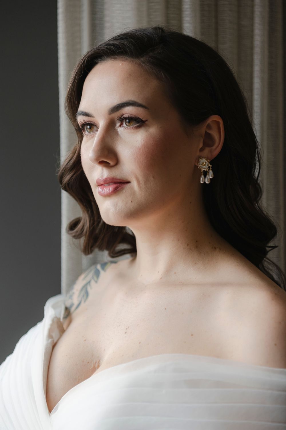 Elegant bridal portrait with soft lighting by a window, featuring delicate drop earrings and an off-shoulder white gown.