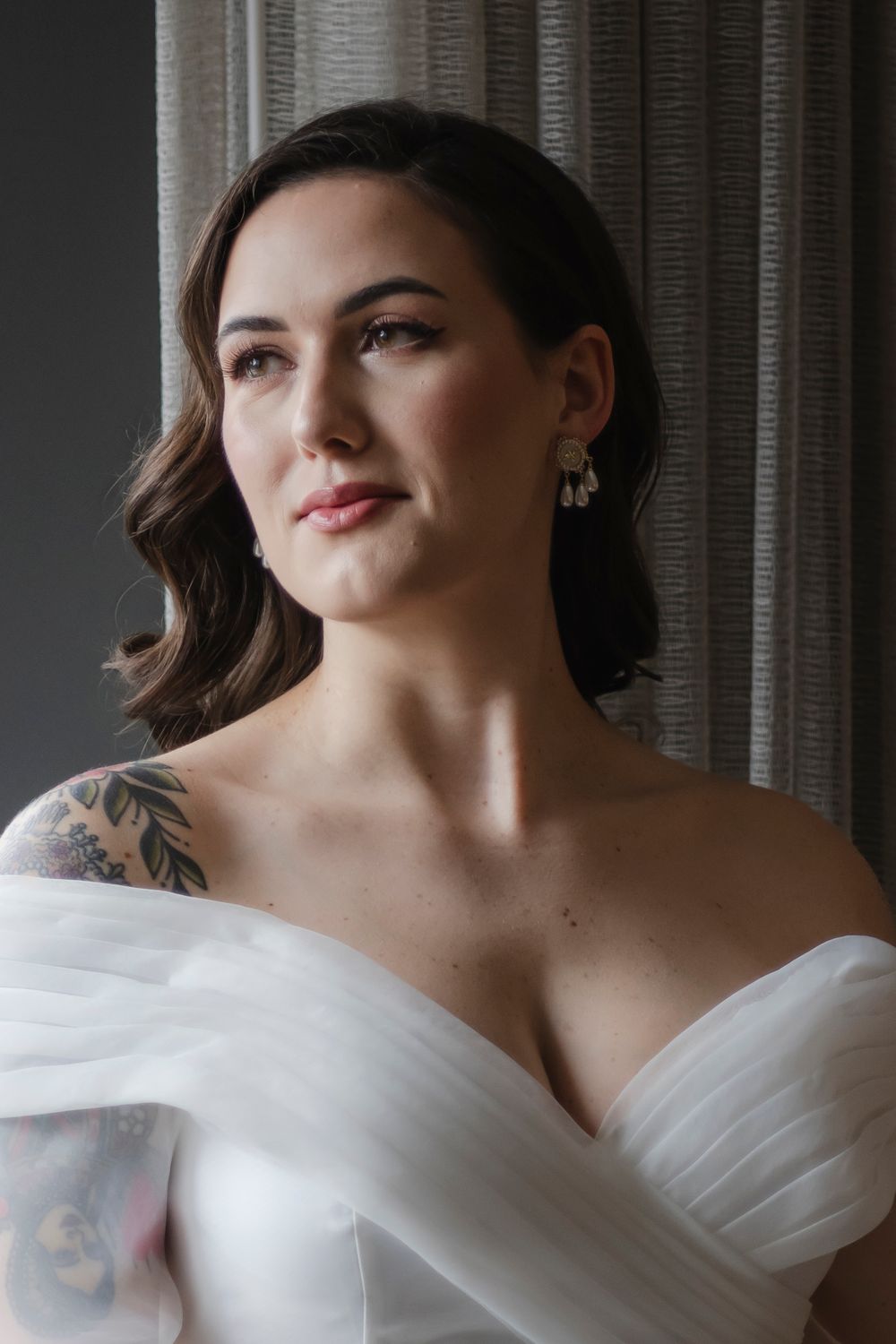 Elegant portrait in soft natural light featuring a white off-shoulder wedding dress and visible shoulder tattoos.