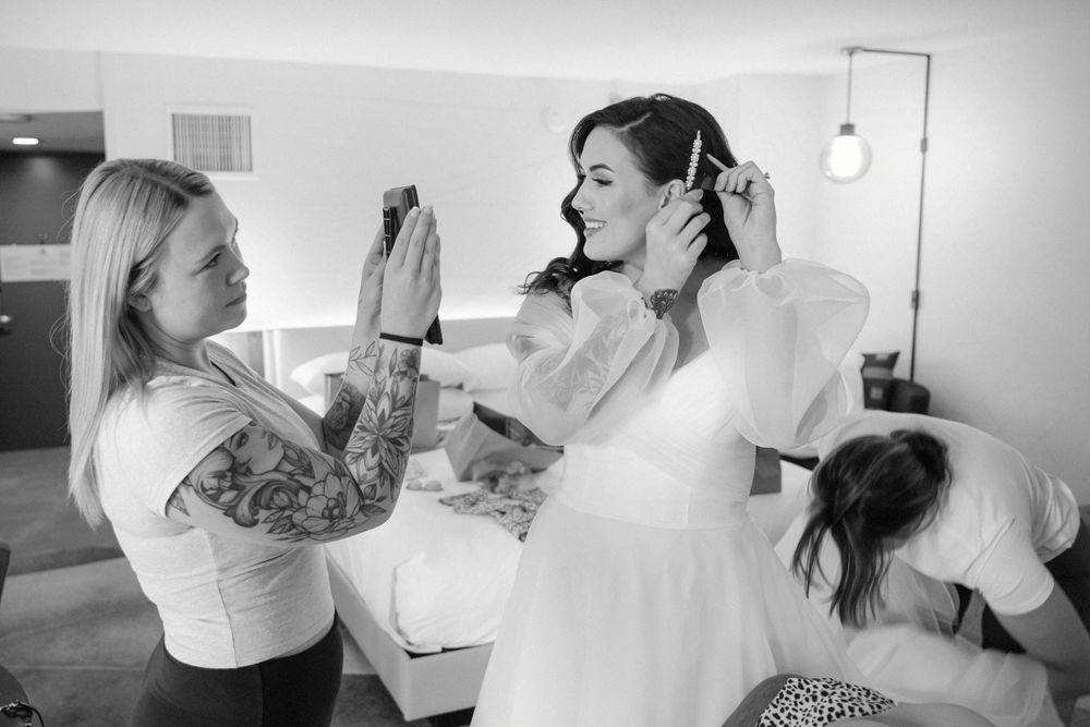 Black and white photo showing bridal preparations with assistants helping adjust a wedding dress during a pre-ceremony moment.