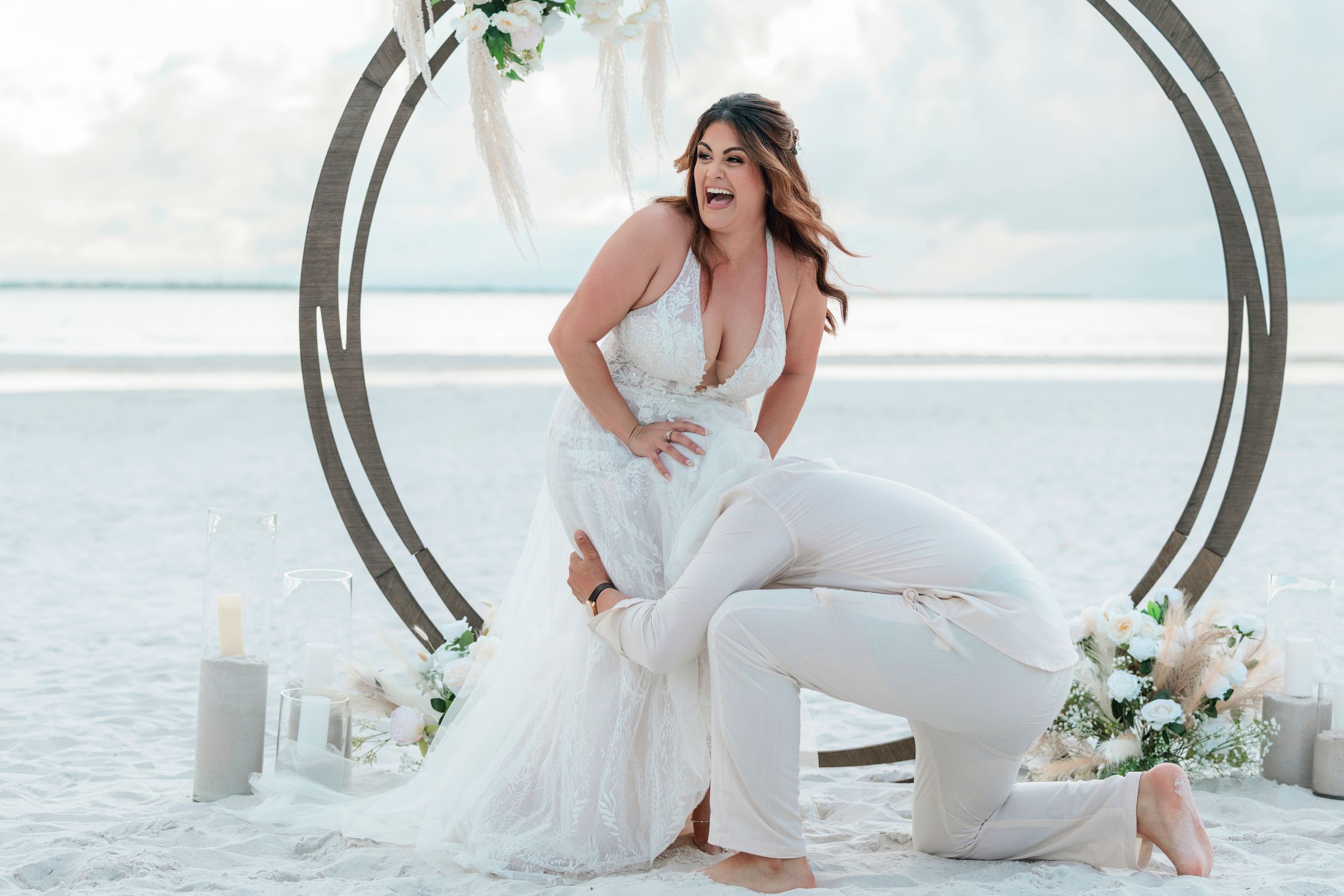 Bride in white lace jumpsuit poses joyfully at beach wedding arch with floral decor.