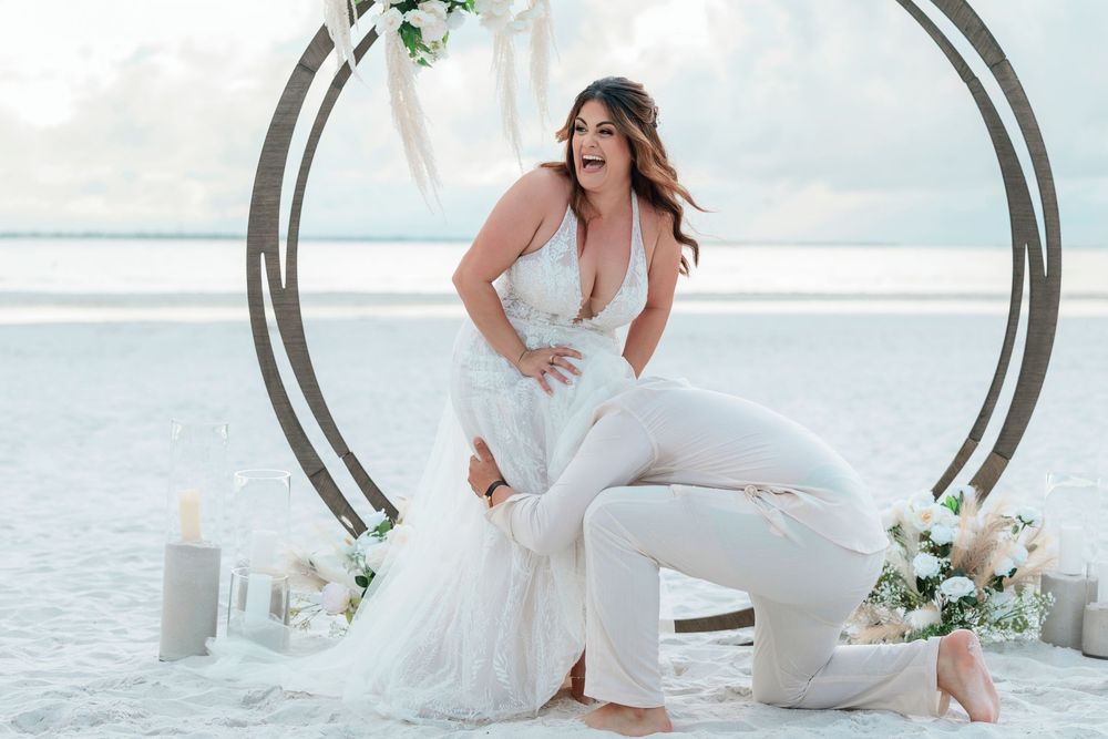 Bride in white lace jumpsuit poses joyfully at beach wedding arch with floral decor.