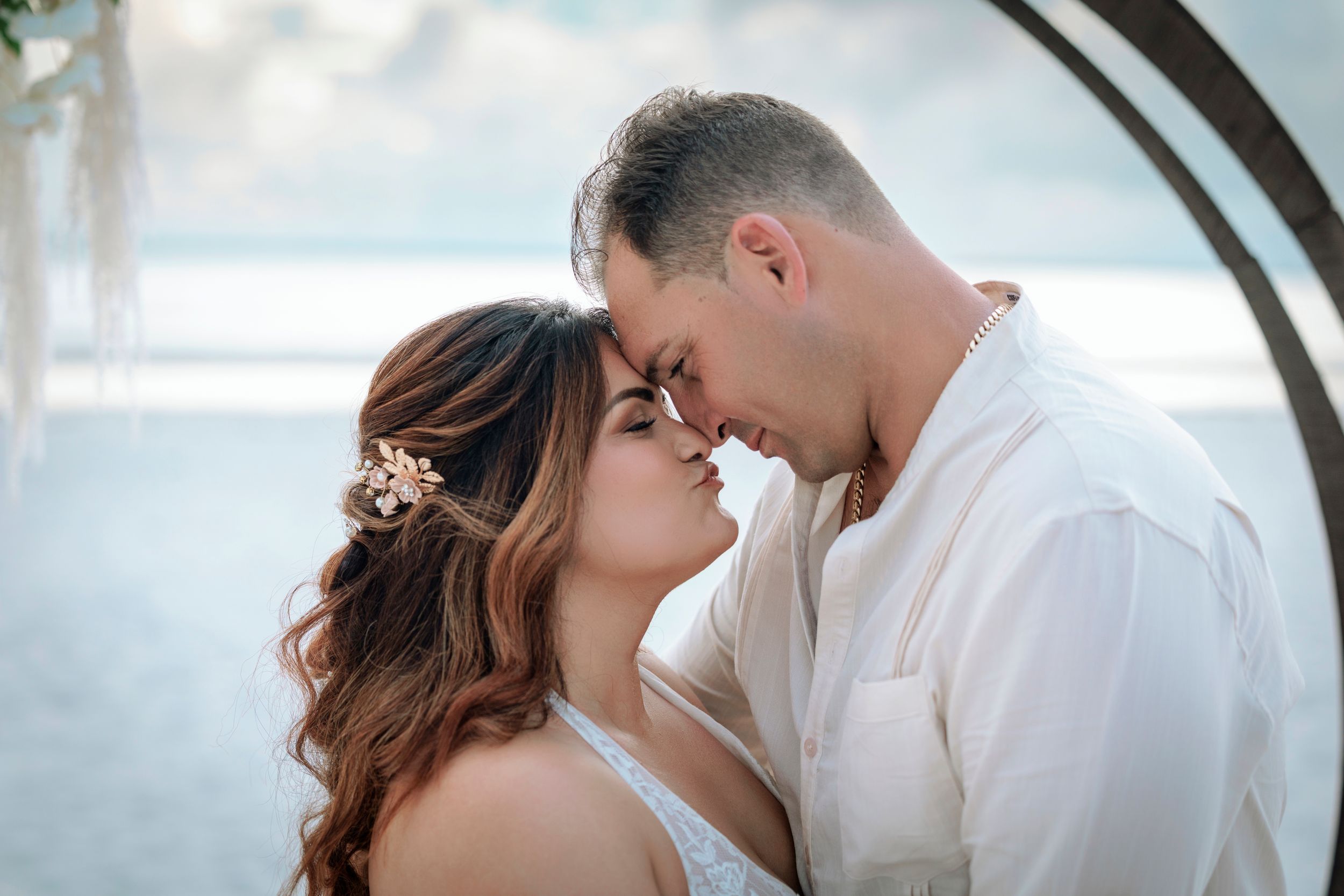 Bride and groom share intimate moment touching foreheads under arch at beach wedding.