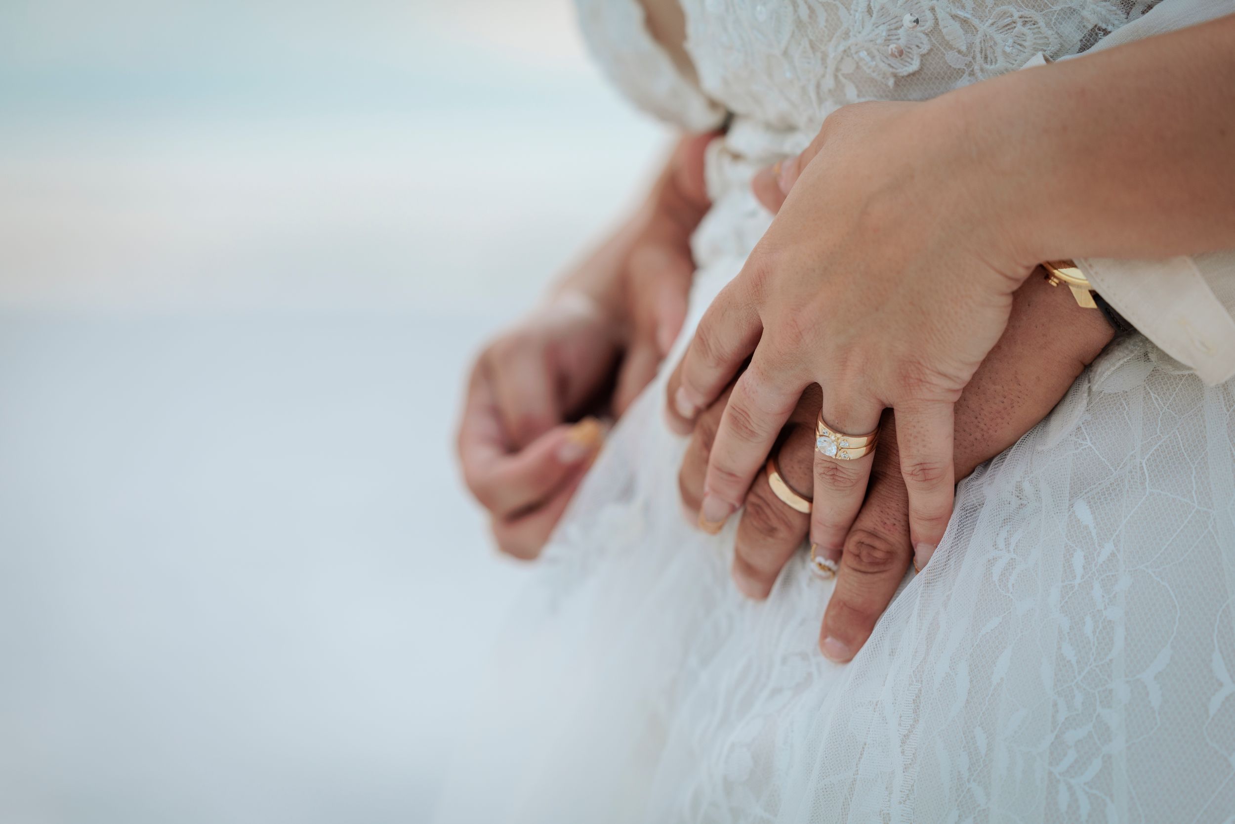 Close-up of newlyweds hands showing gold wedding bands on white lace wedding dress.