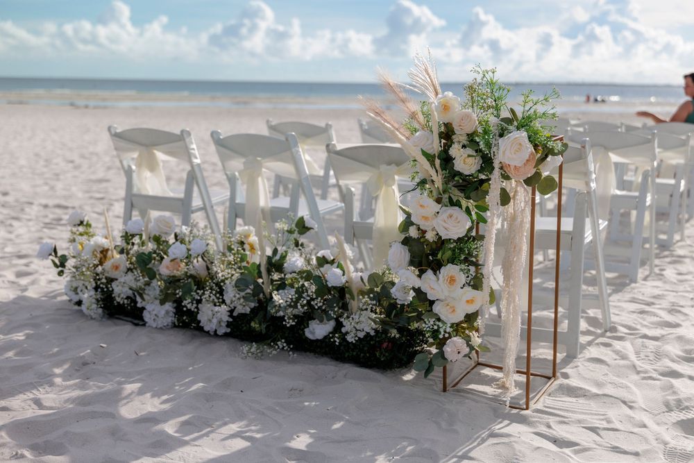 Elegant beach wedding ceremony setup with white chairs and white floral arrangements on sand.