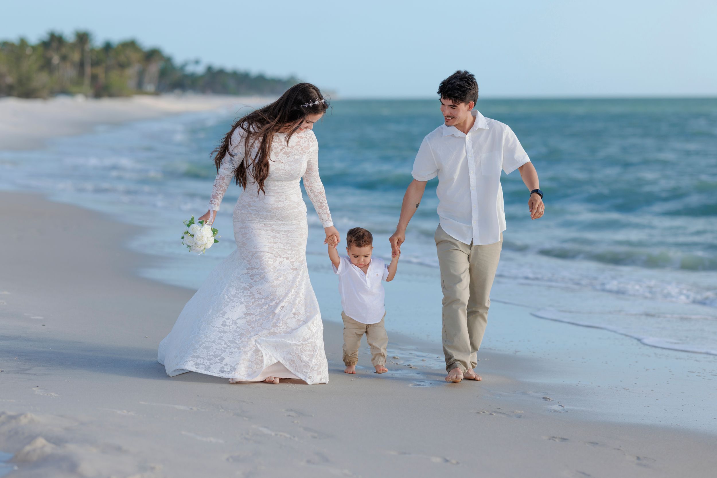 Wedding couple walks on beach with toddler son, bride in white lace gown holding bouquet.