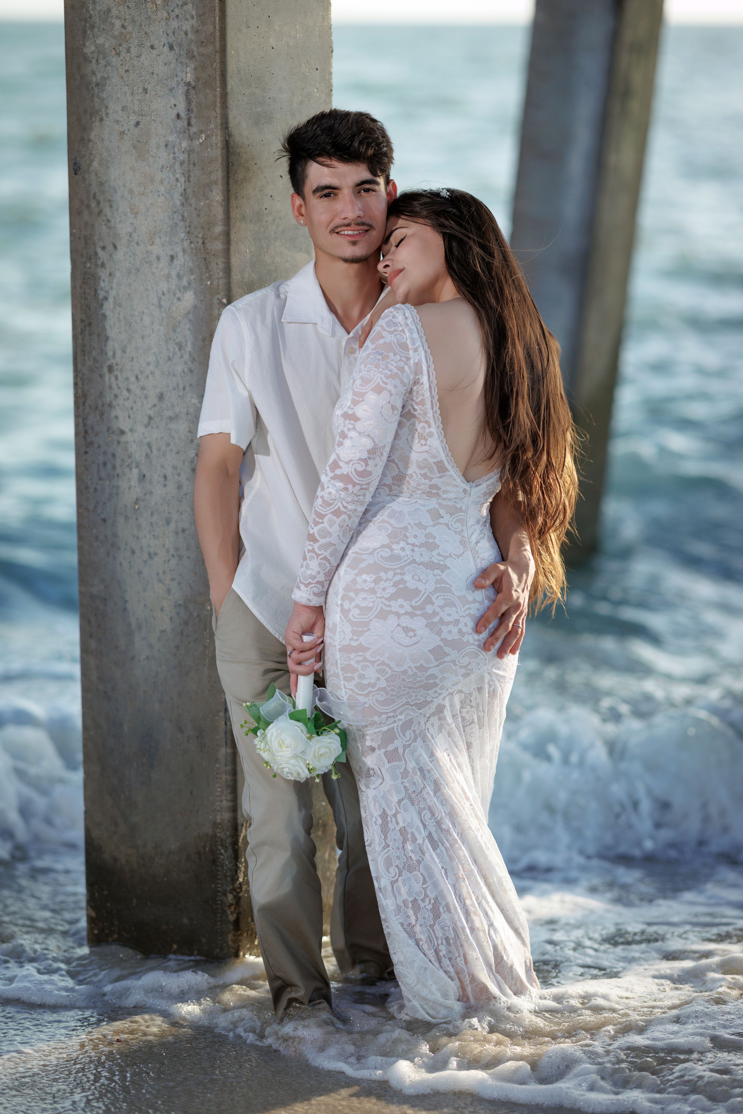 Bride in backless lace gown and groom pose by pier pillar with ocean waves.