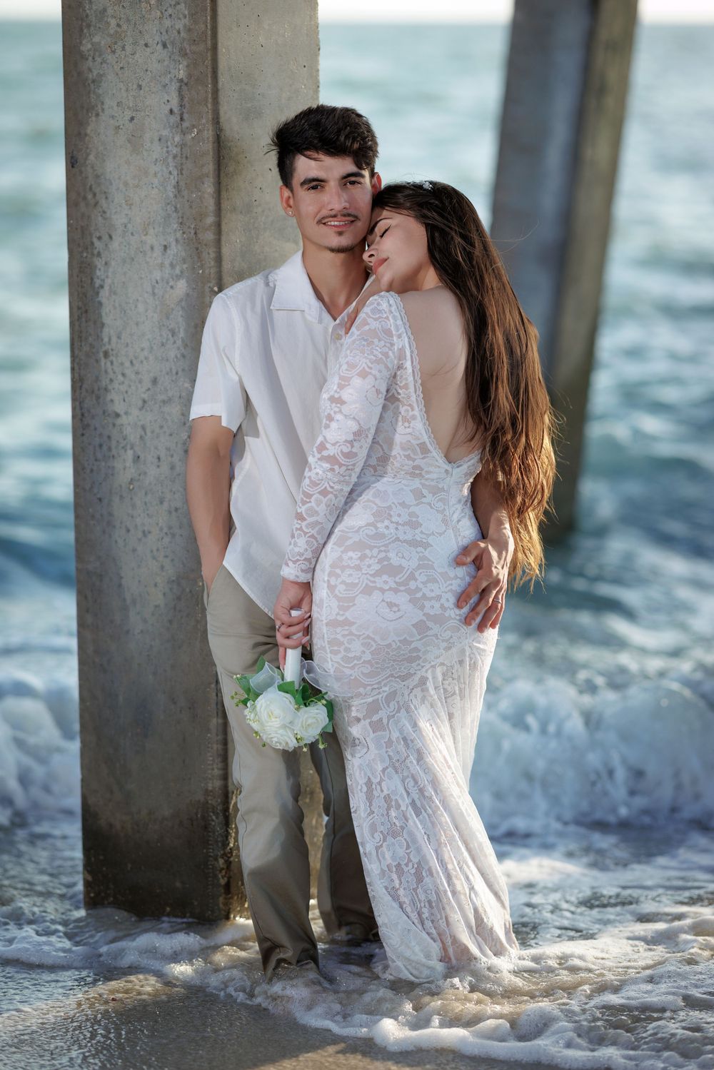 Bride in backless lace gown and groom pose by pier pillar with ocean waves.
