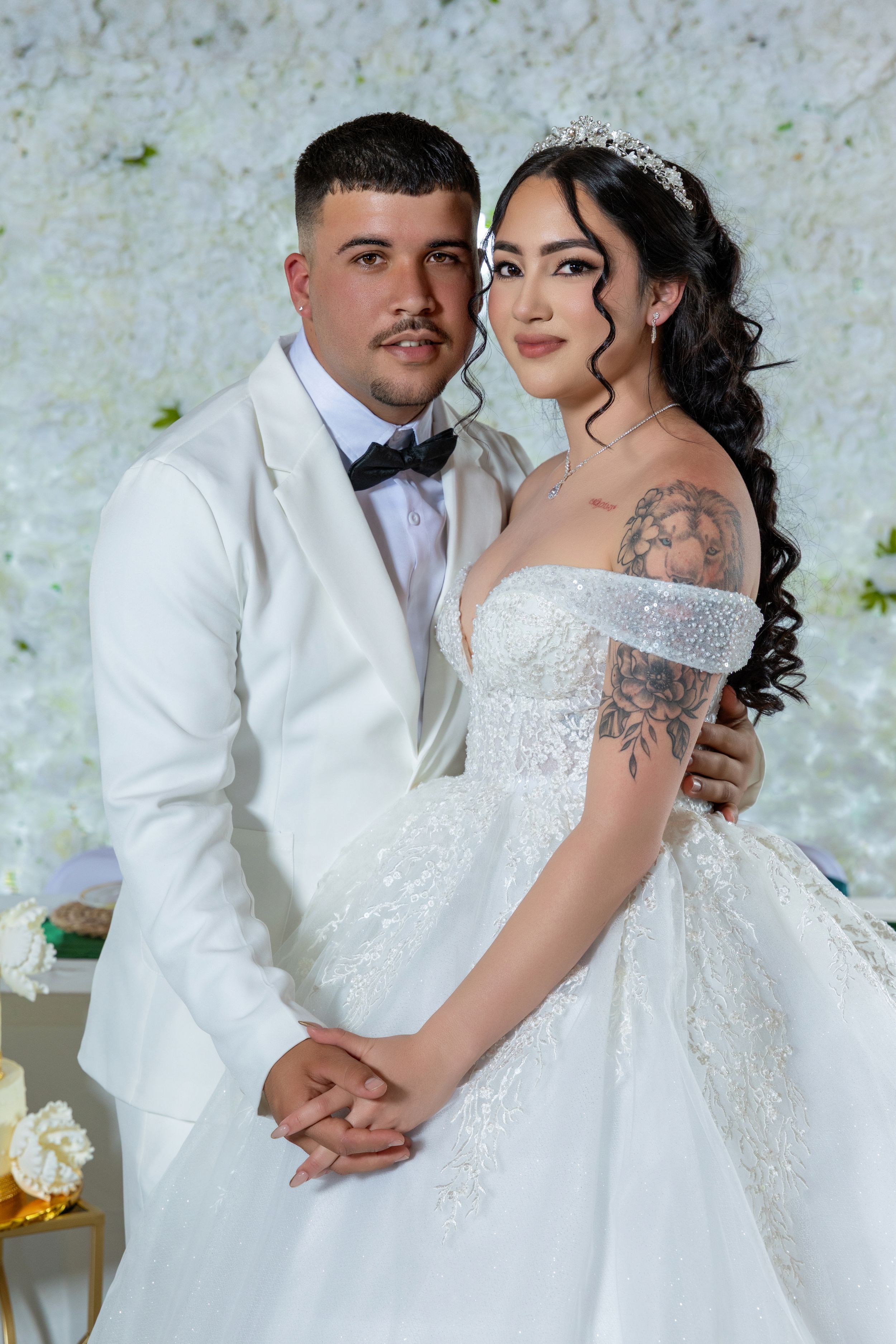 Elegant couple in white wedding attire pose before white floral wall backdrop.