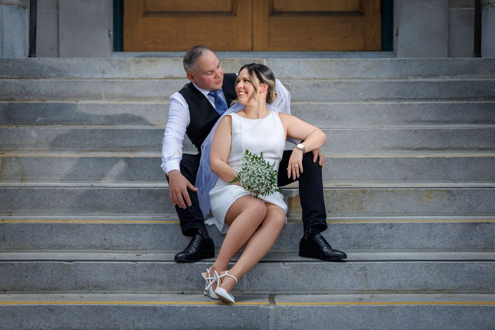 Happy couple in wedding attire sitting on stone steps, bride holding white bouquet in short white dress with veil.