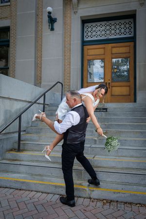 Groom carries laughing bride over shoulder on steps outside historic brick building.