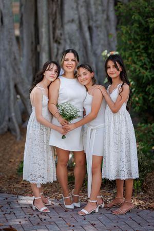 Bride in white mini dress poses with three young girls in white lace dresses outdoors.