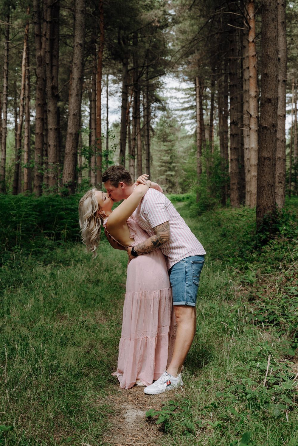 A romantic couple embraces on a forest path surrounded by tall pine trees and lush greenery.