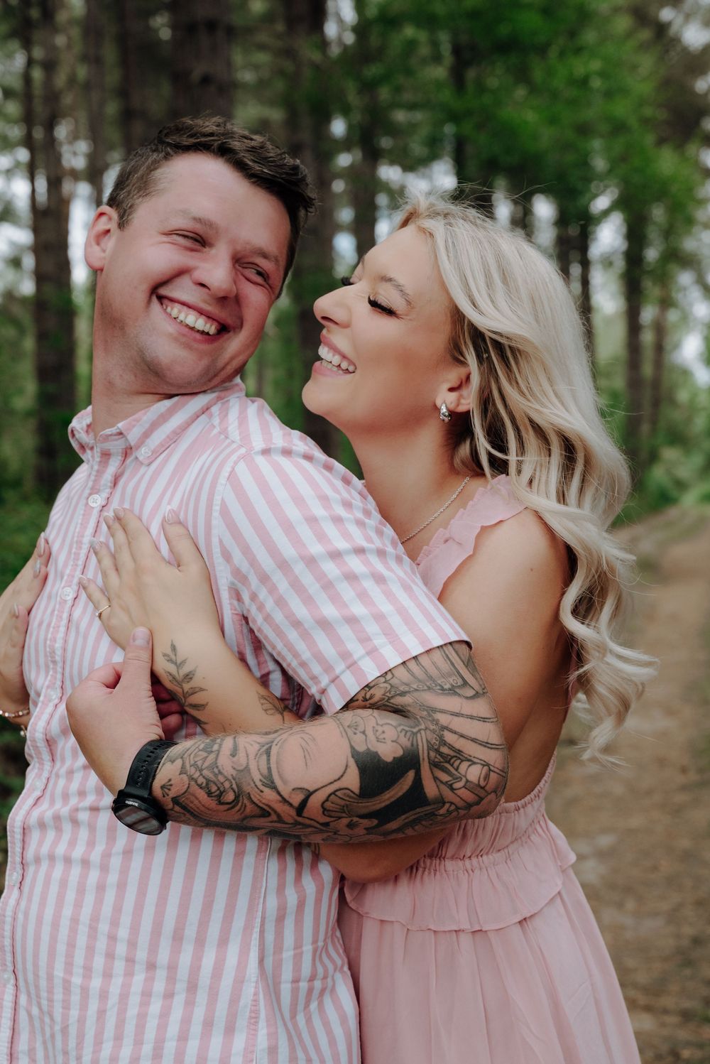A couple in pink outfits embraces and laughs together during a romantic photoshoot in a wooded setting.