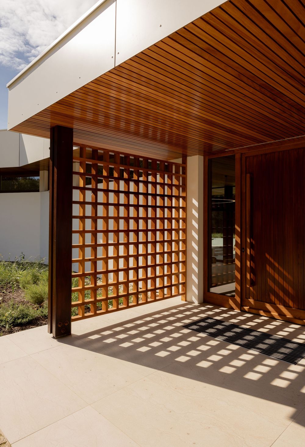 Modern wooden lattice structure creates mesmerizing shadow patterns on white concrete in outdoor architectural space.