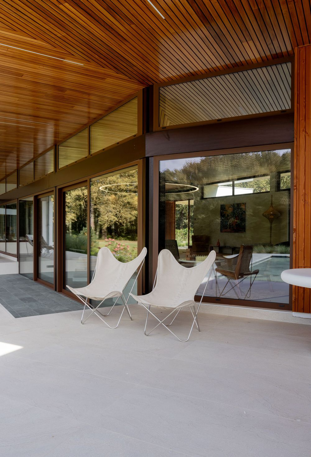 Modern covered patio with wood ceiling features sleek white outdoor chairs and panoramic views through floor-to-ceiling windows.