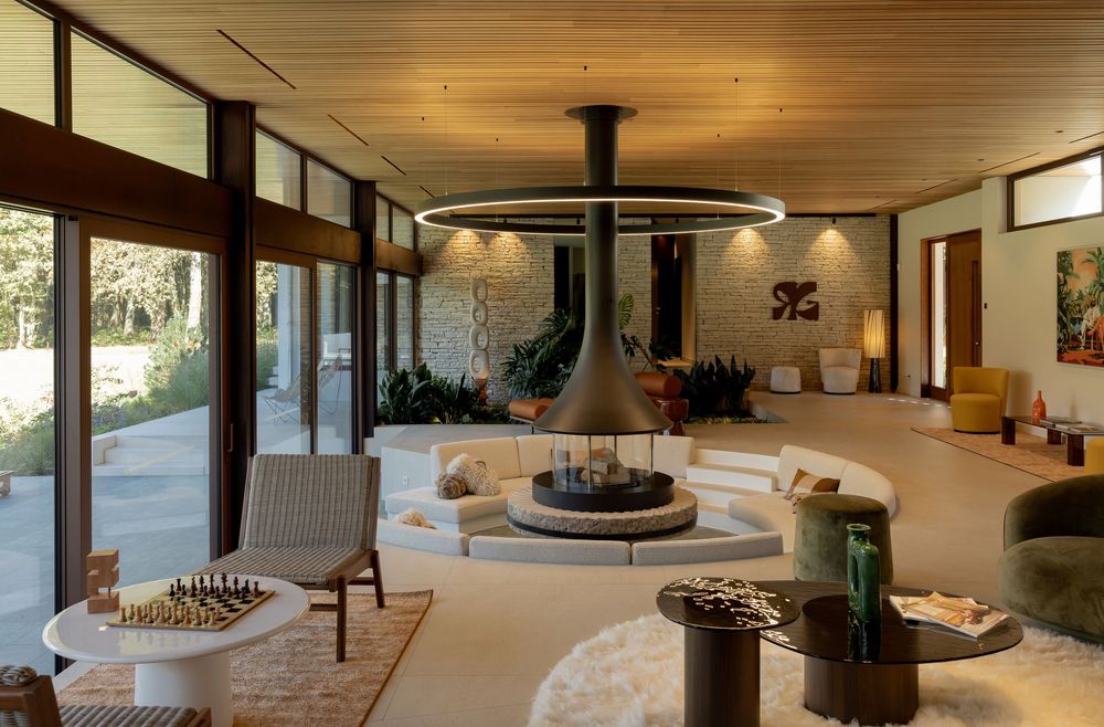 Modern open concept living room with wood ceiling, hanging fireplace, and floor-to-ceiling windows overlooking nature.