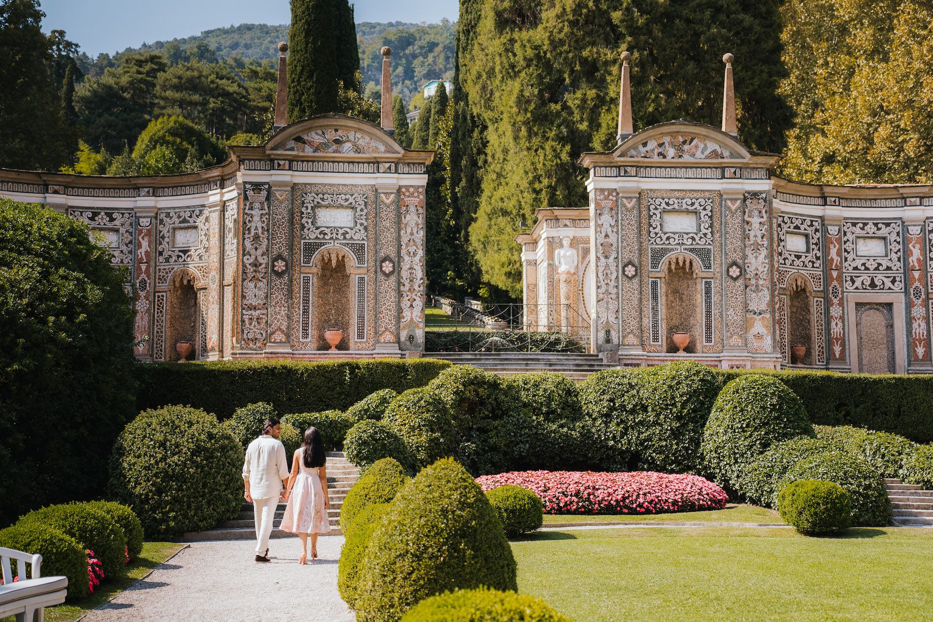 Scenic garden walkway with ornate classical architecture, trimmed hedges, and colorful flower beds in a grand estate setting.