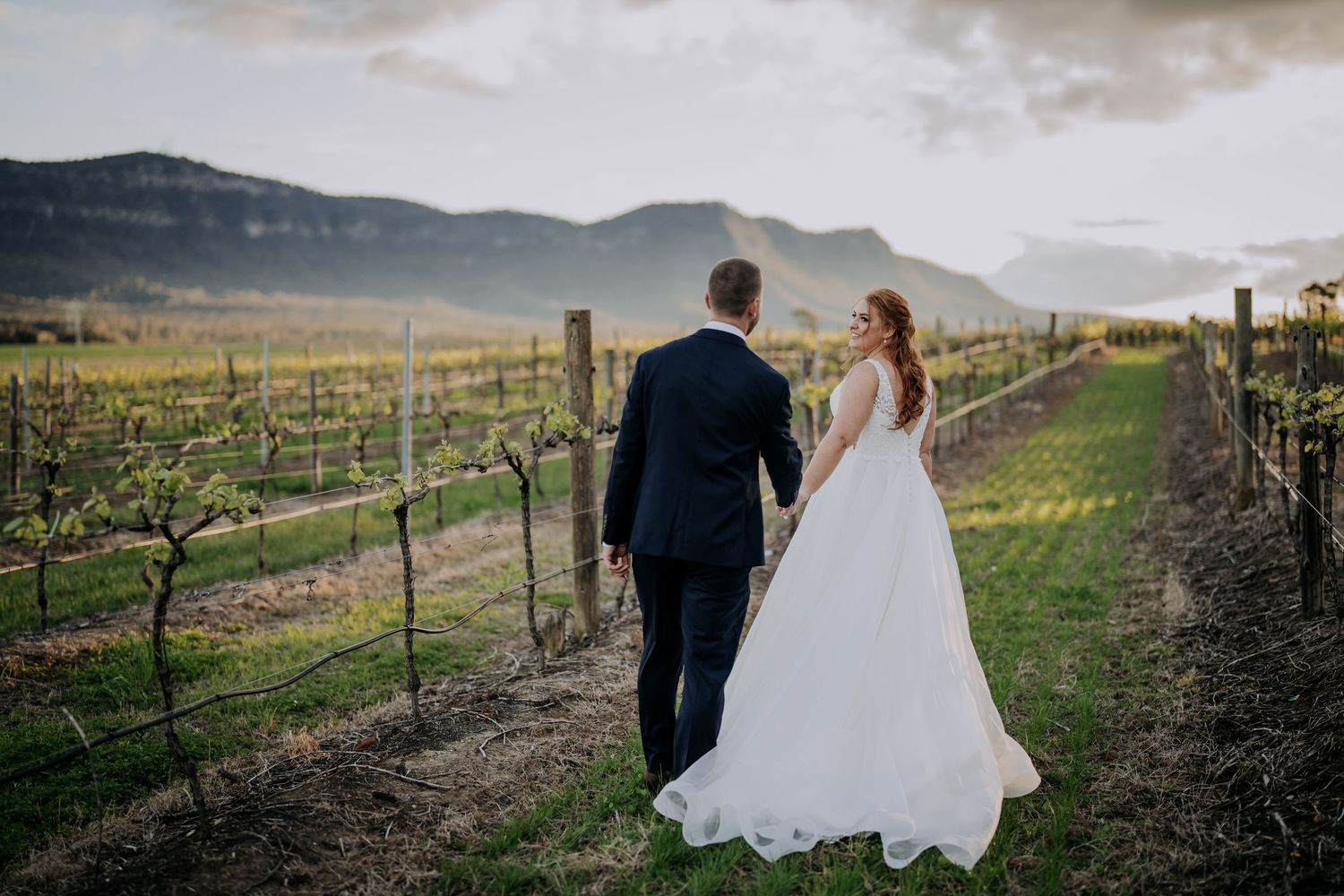 Bride and groom walking through vineyard with mountain landscape in background.