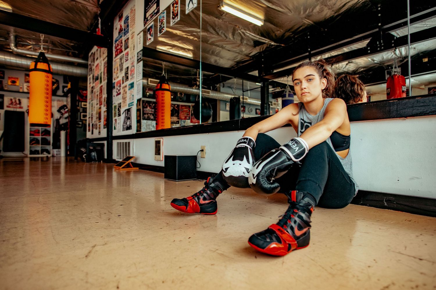 Female boxer in boxing gloves sitting on gym floor in athletic wear with red and black sneakers.