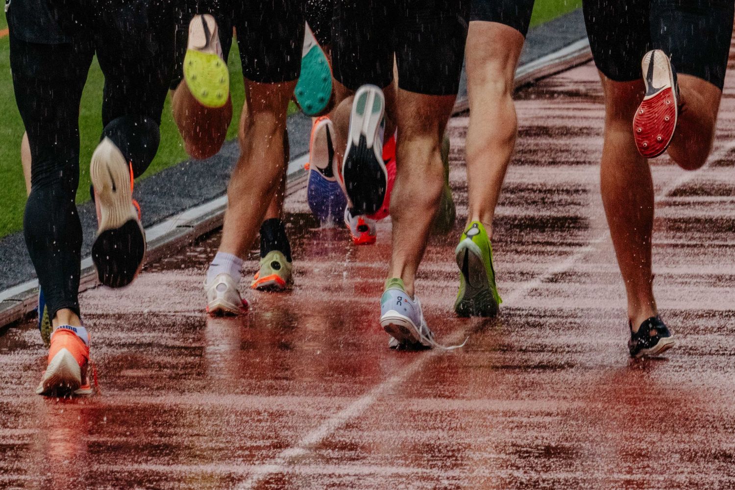 Athletes sprint in rain on wet track wearing colorful running spikes during competitive track meet.