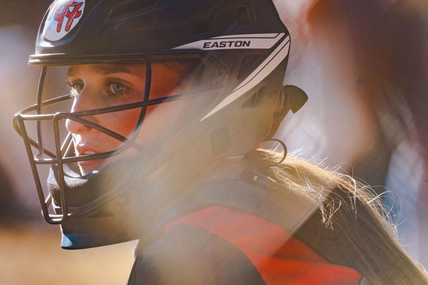 Close-up of female softball player wearing Easton helmet with face guard, hair flowing in warm sunlight.