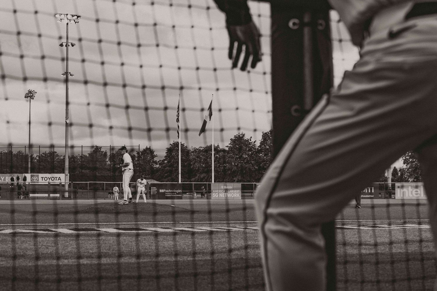 Black and white view through baseball backstop netting of pitcher on mound during game.