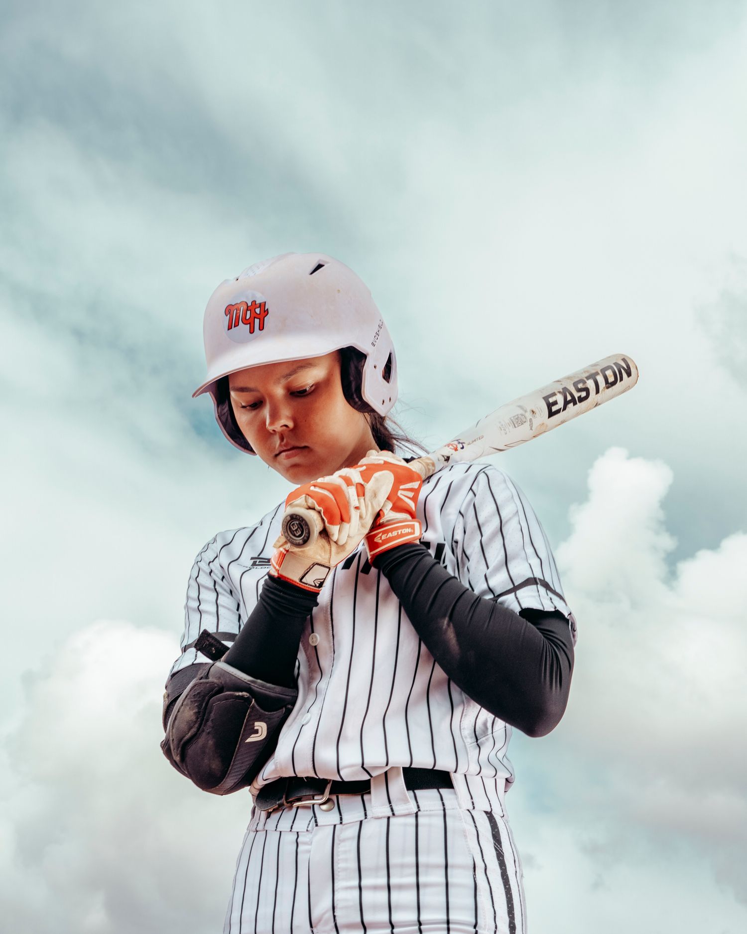 Young softball player in pinstripe uniform grips Easton bat, wearing helmet and batting gloves under cloudy sky.