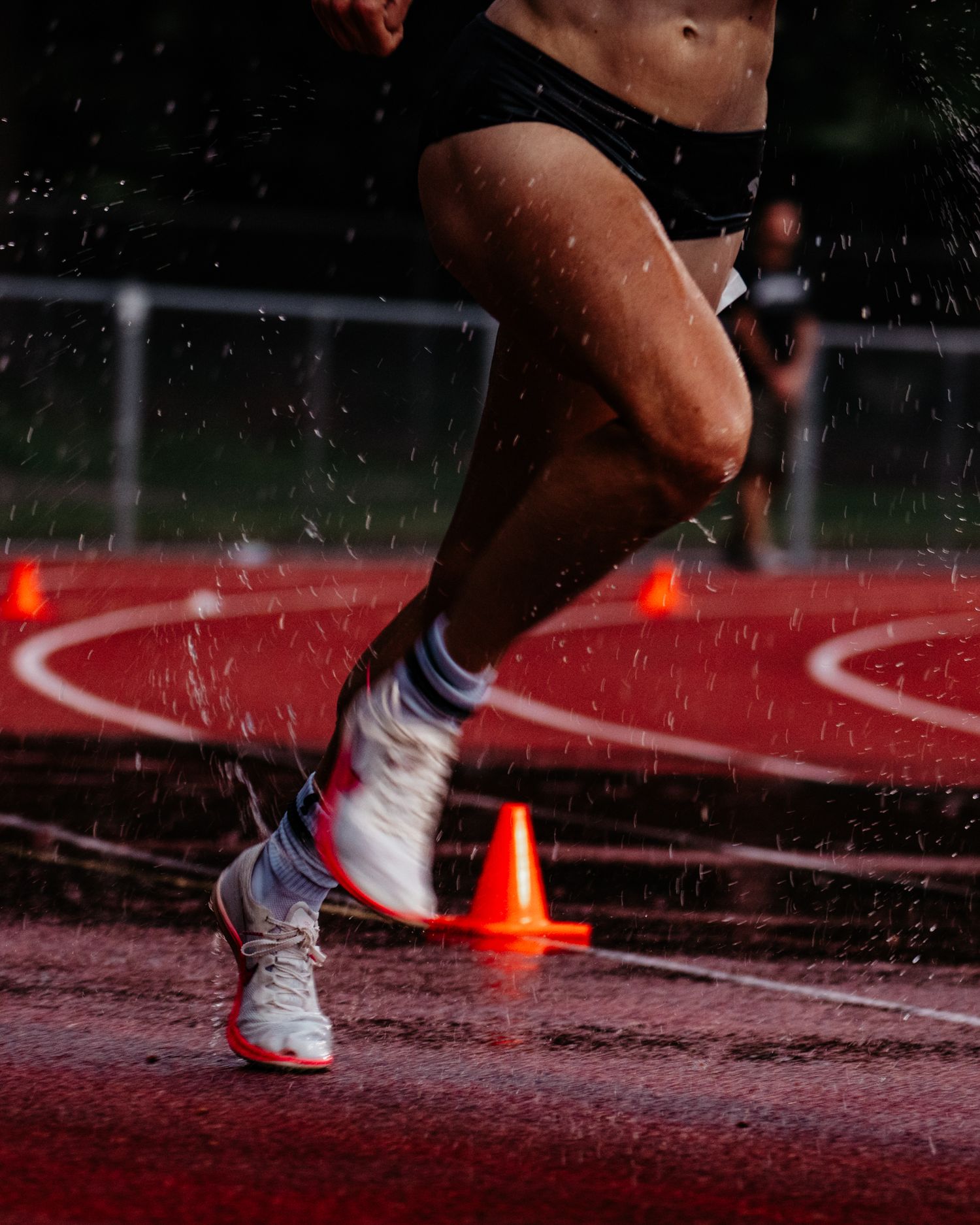 Female runner sprints past orange cone on wet red track in rain, wearing white athletic shoes.