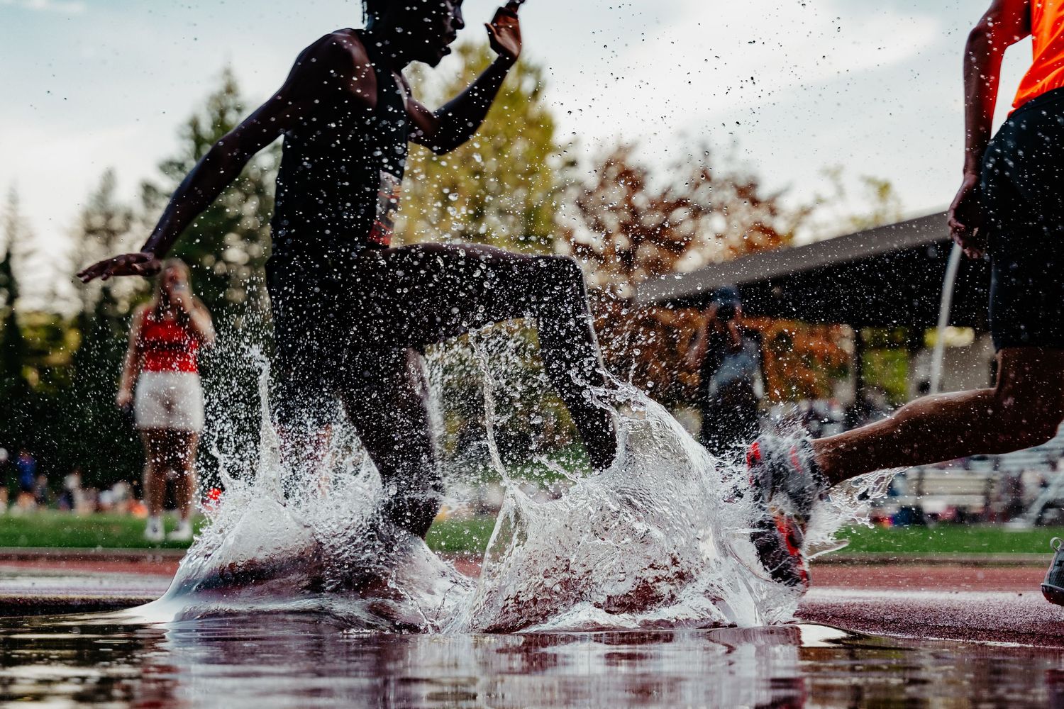 Track athletes splash through steeplechase water pit during outdoor competition in dramatic action shot.