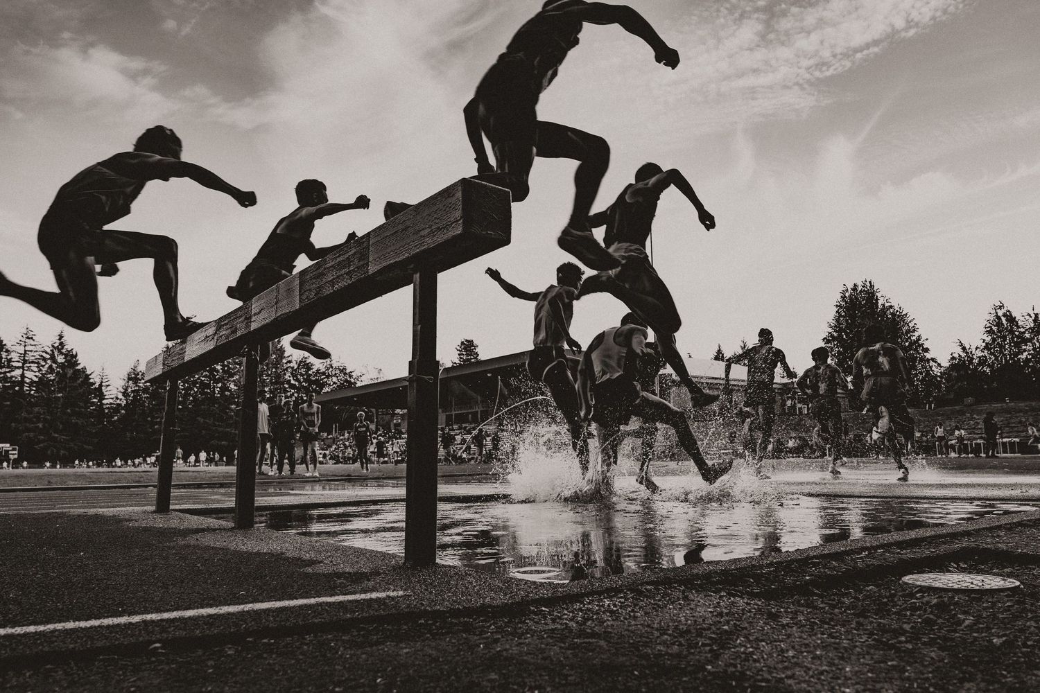 Black and white photo of steeplechase runners leaping over water barrier during track competition.