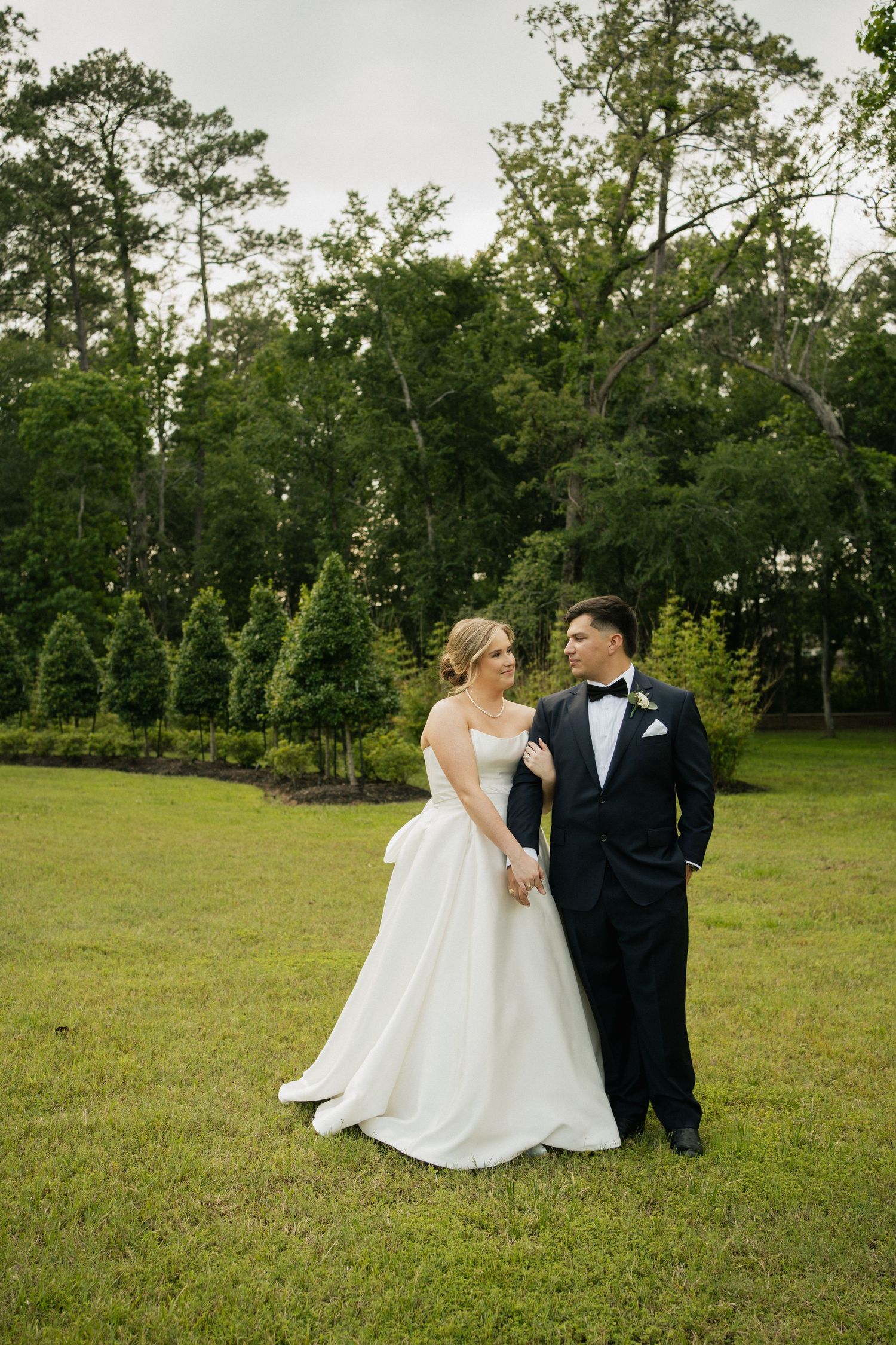 A couple poses in formal wedding attire on a manicured lawn with pine trees in the background.