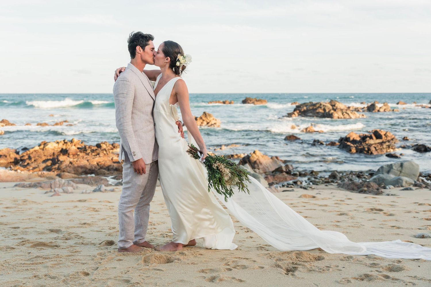 Romantic beach wedding portrait with couple standing on rocky coastline at sunset.